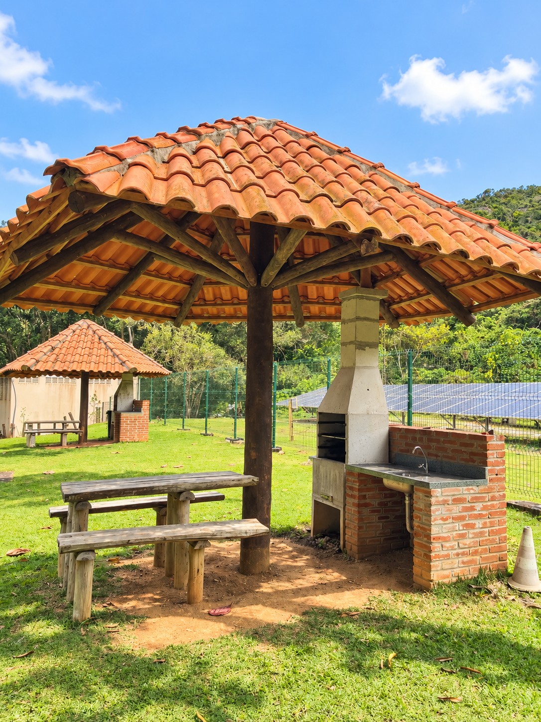 Quiosque com telhado de telha, mesa com bancos e churrasqueira ao fundo